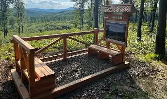 Drewniana infrastruktura turystyczna z ławkami i tablicą informacyjną ustawiona w lesie z widokiem na góry