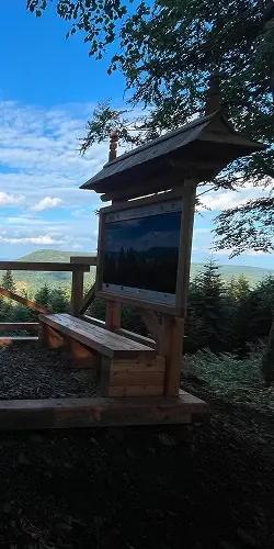 Drewniana ławka z tablicą informacyjną na punkcie widokowym w Beskidzie Niskim, z panoramą gór w tle.